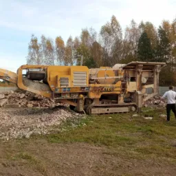 Ż&oacute;łty mobilny kruszarka EXTEC na gąsienicach, obok sterta gruzu. W tle drzewa i betonowe ogrodzenie. Mężczyzna w białej koszulce stoi obok maszyny.