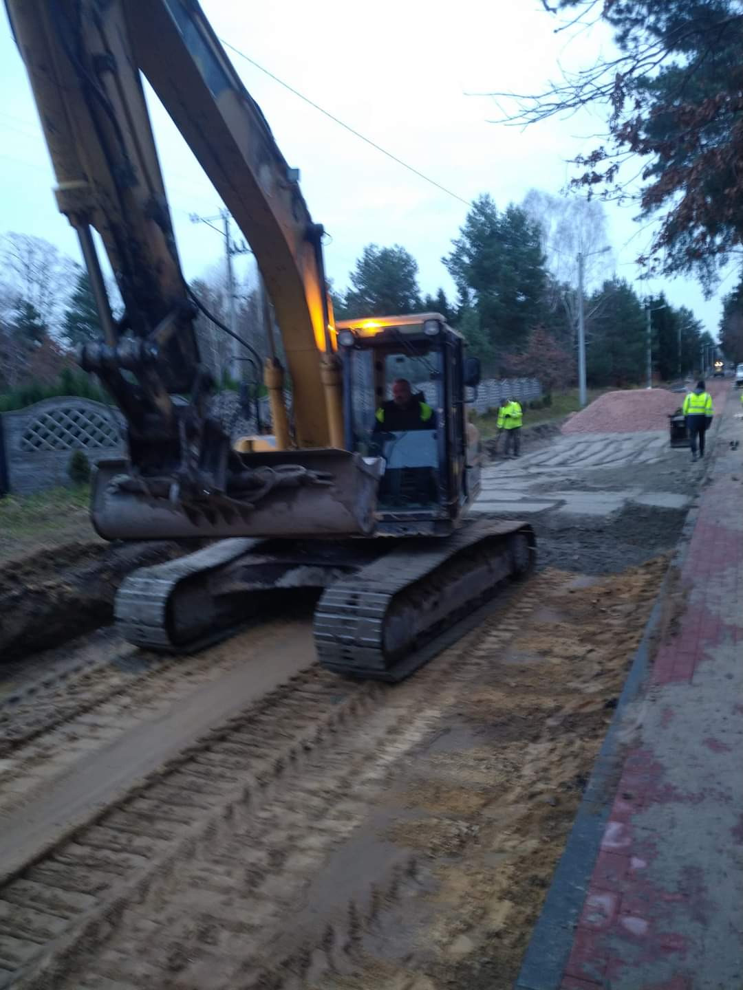 Żółta koparka gąsienicowa w trakcie prac ziemnych przy drodze, operator w kabinie, pracownicy w odblaskowych kamizelkach w tle. Etap utwardzania drogi.