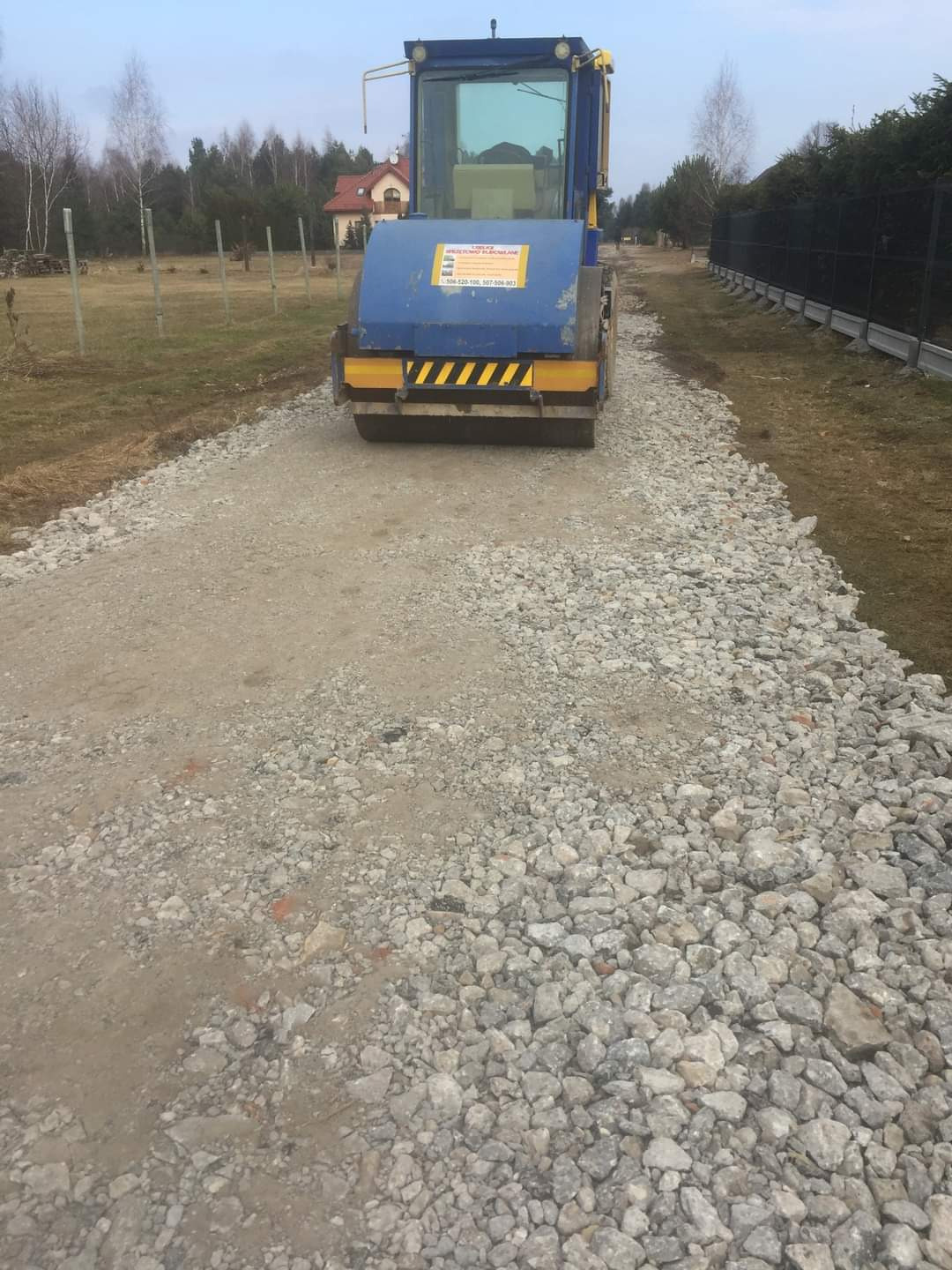 Walec drogowy ubija kamienistą nawierzchnię drogi gruntowej na tle zieleni i ogrodzenia. Widoczne ślady ubijania, niebieski walec z żółtymi akcentami.