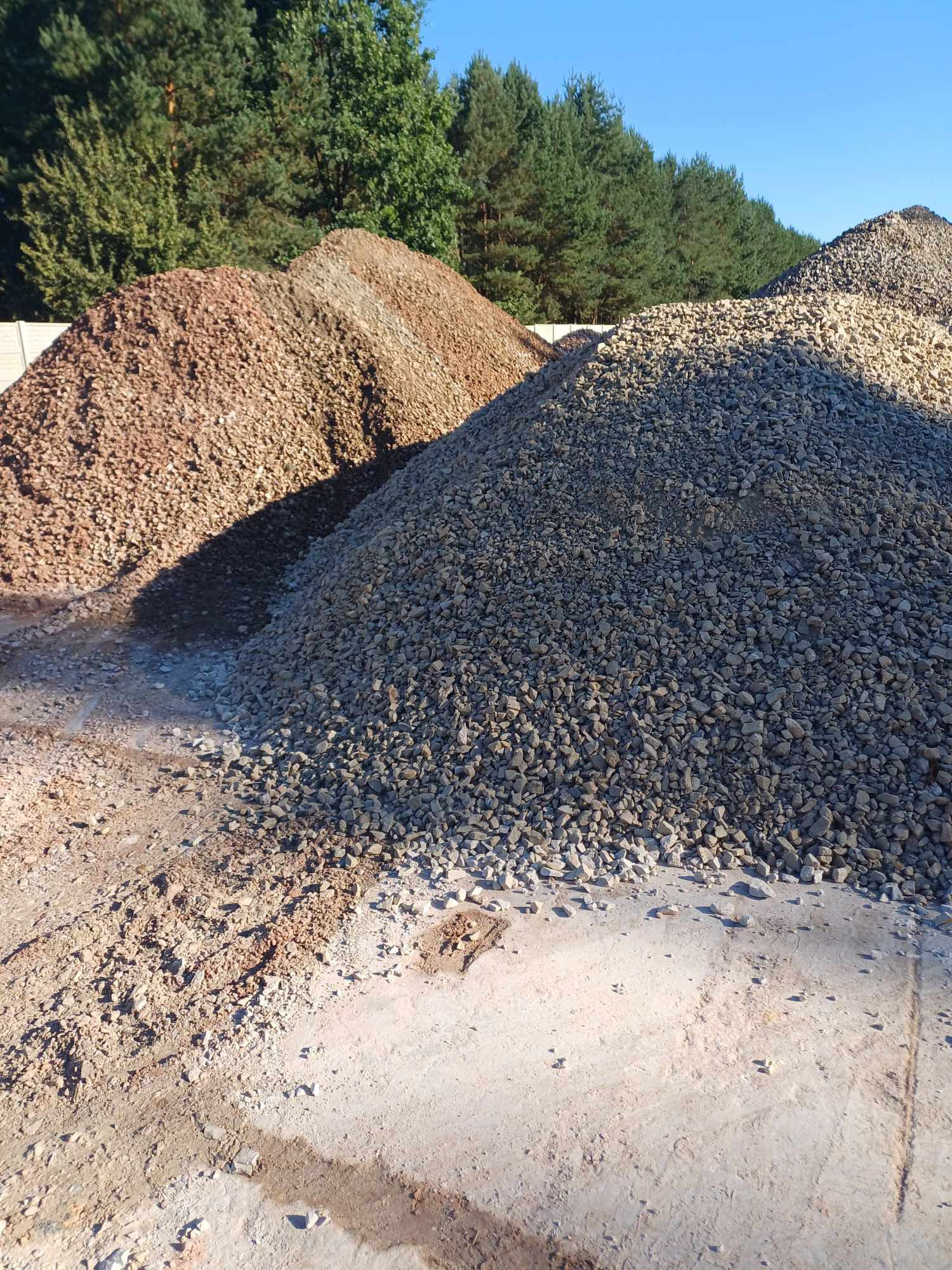 Góra szarego gruzu na piaskowym podłożu, za nią wzniesienia z czerwonego i beżowego gruzu. W tle drzewa i błękitne niebo.