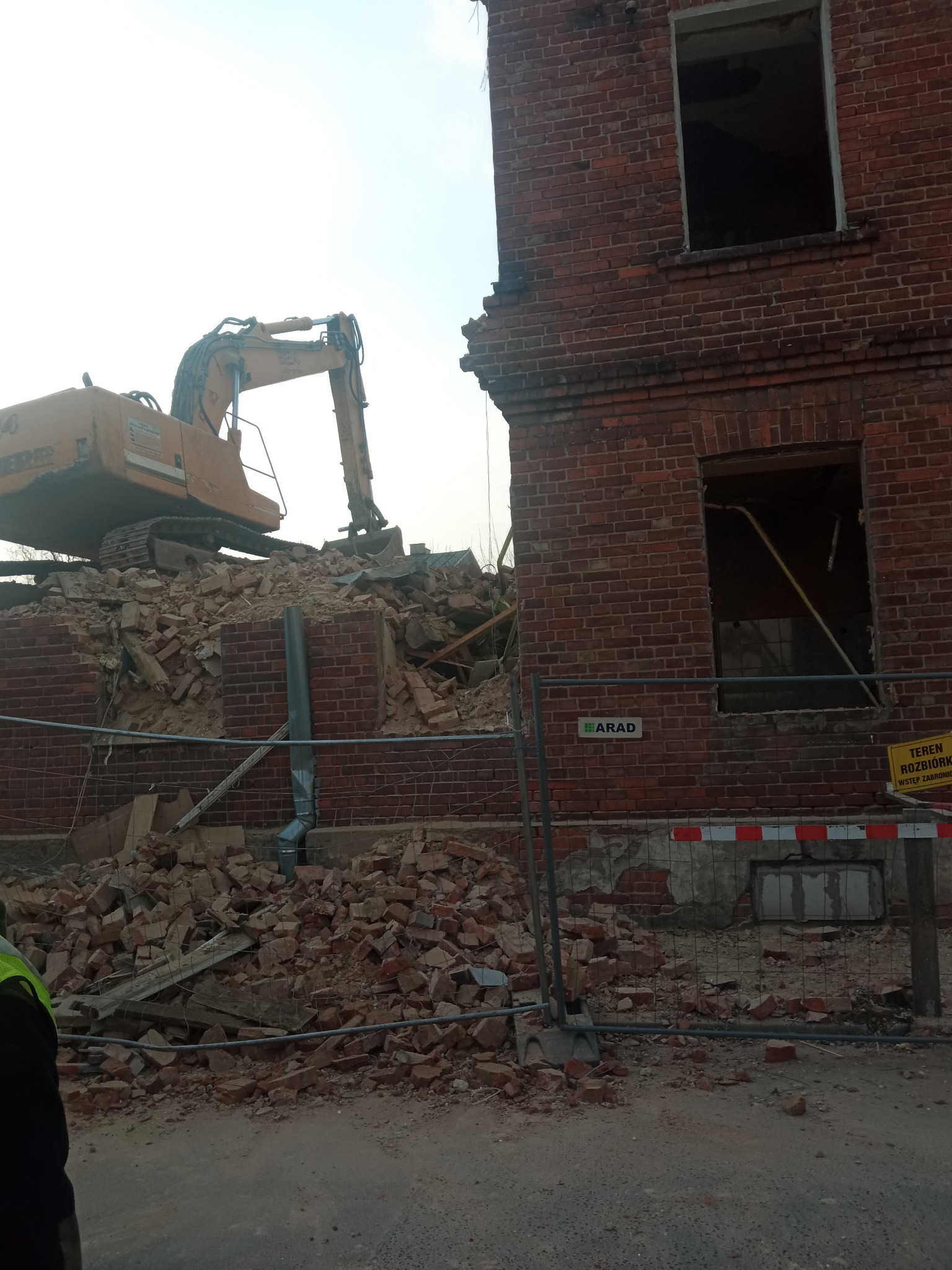 Częściowo zburzony, ceglany budynek z widocznym sprzętem budowlanym na gruzowisku. Żółta tablica ostrzegawcza przy ogrodzeniu. Widok z dołu.