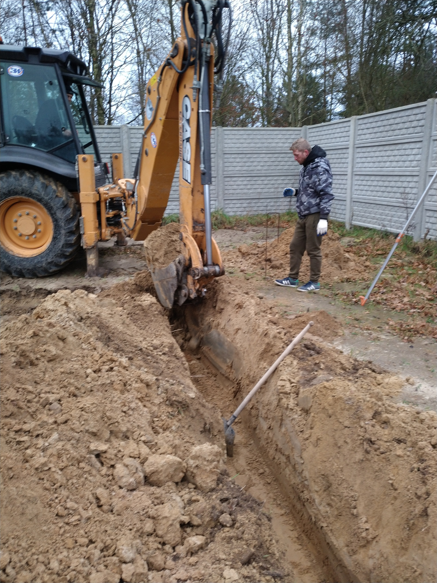 Żółta koparko-ładowarka w trakcie wykopu wąskiego rowu w ziemi. Obok stoi pracownik w rękawicach, kontrolujący postęp prac. W tle betonowe ogrodzenie.