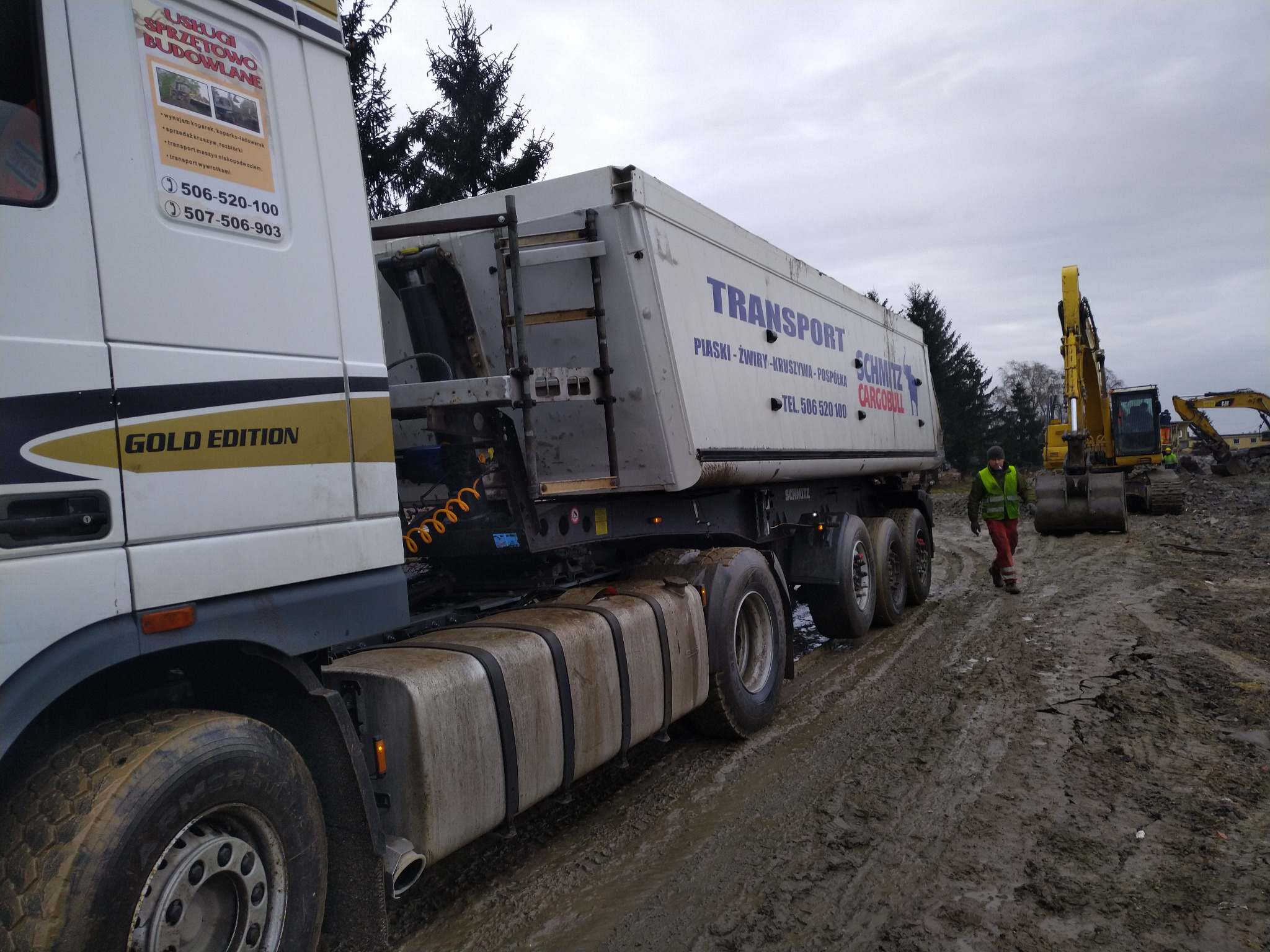 Ciężarówka z naczepą Schmitz CargoBull na błotnistym terenie budowy, w tle koparki i pracownik w kamizelce odblaskowej. Widoczne reklamy usług transportowych i budowlanych.