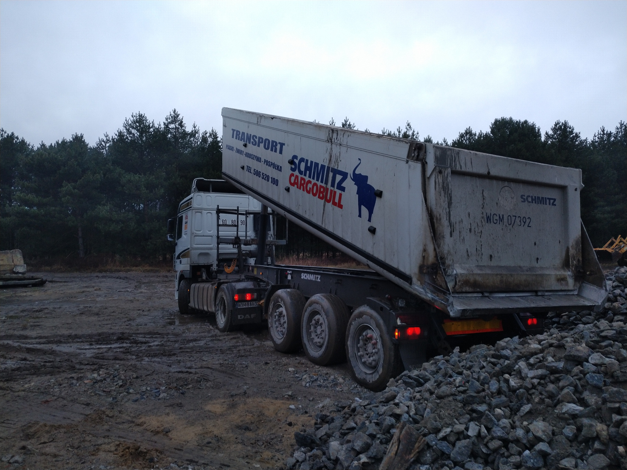 Ciężarówka wywrotka Schmitz CargoBull z podniesioną skrzynią ładunkową, wysypująca gruz na placu budowy. W tle las iglasty i pochmurne niebo.