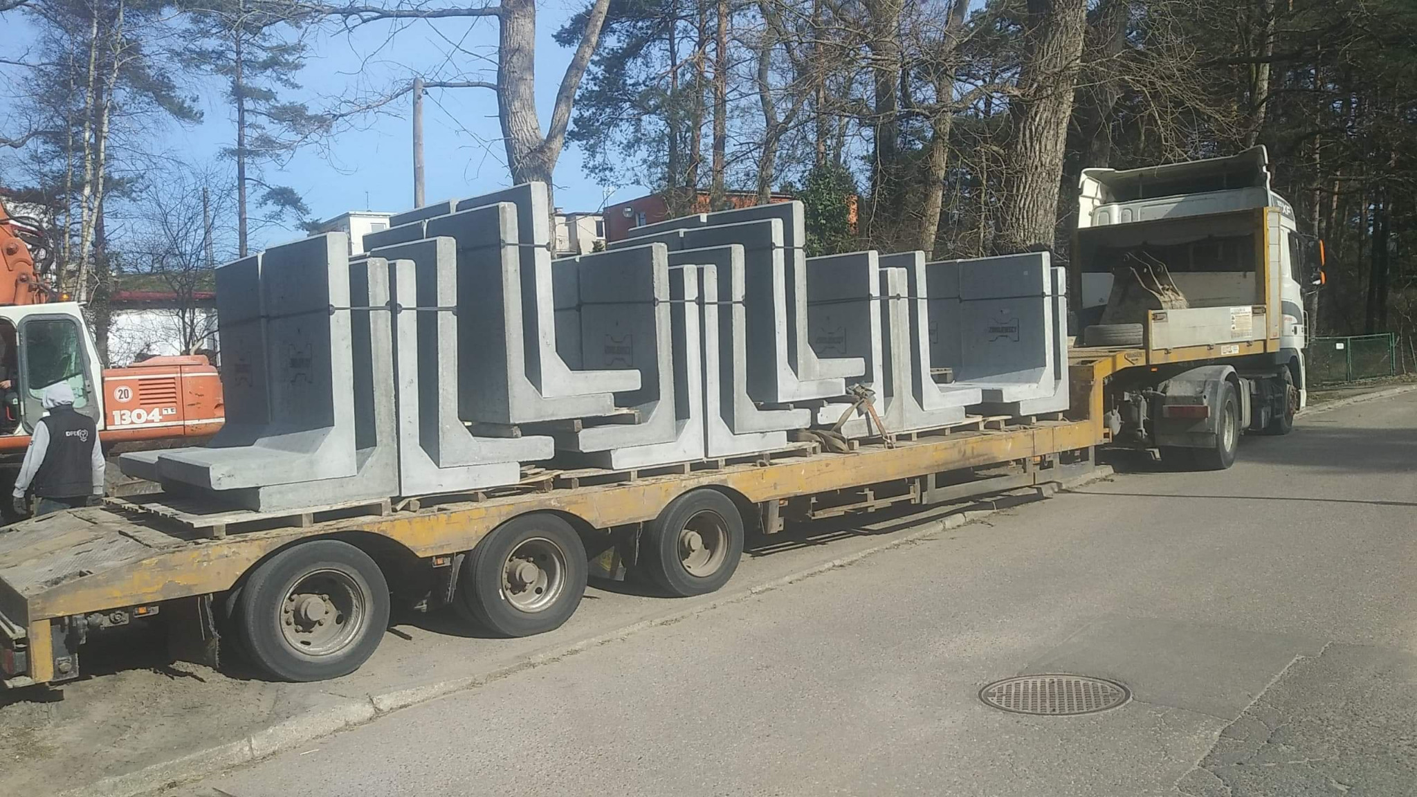 Ciężarówka transportuje betonowe elementy L na lawecie, w tle drzewa i budynek. Widoczny dźwig i pracownik. Transport elementów prefabrykowanych.