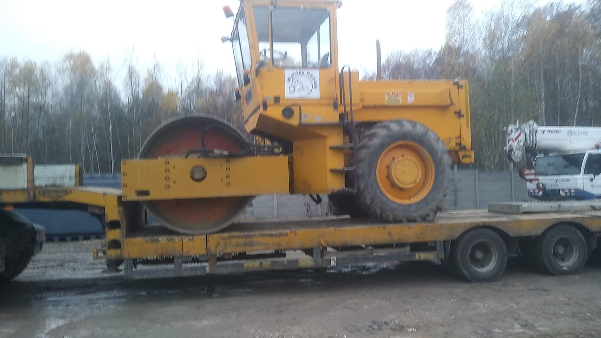 Żółty walec drogowy na niskopodwoziowej platformie transportowej. Widoczne opony, kabina i fragment lasu w tle. Maszyna gotowa do transportu na plac budowy.