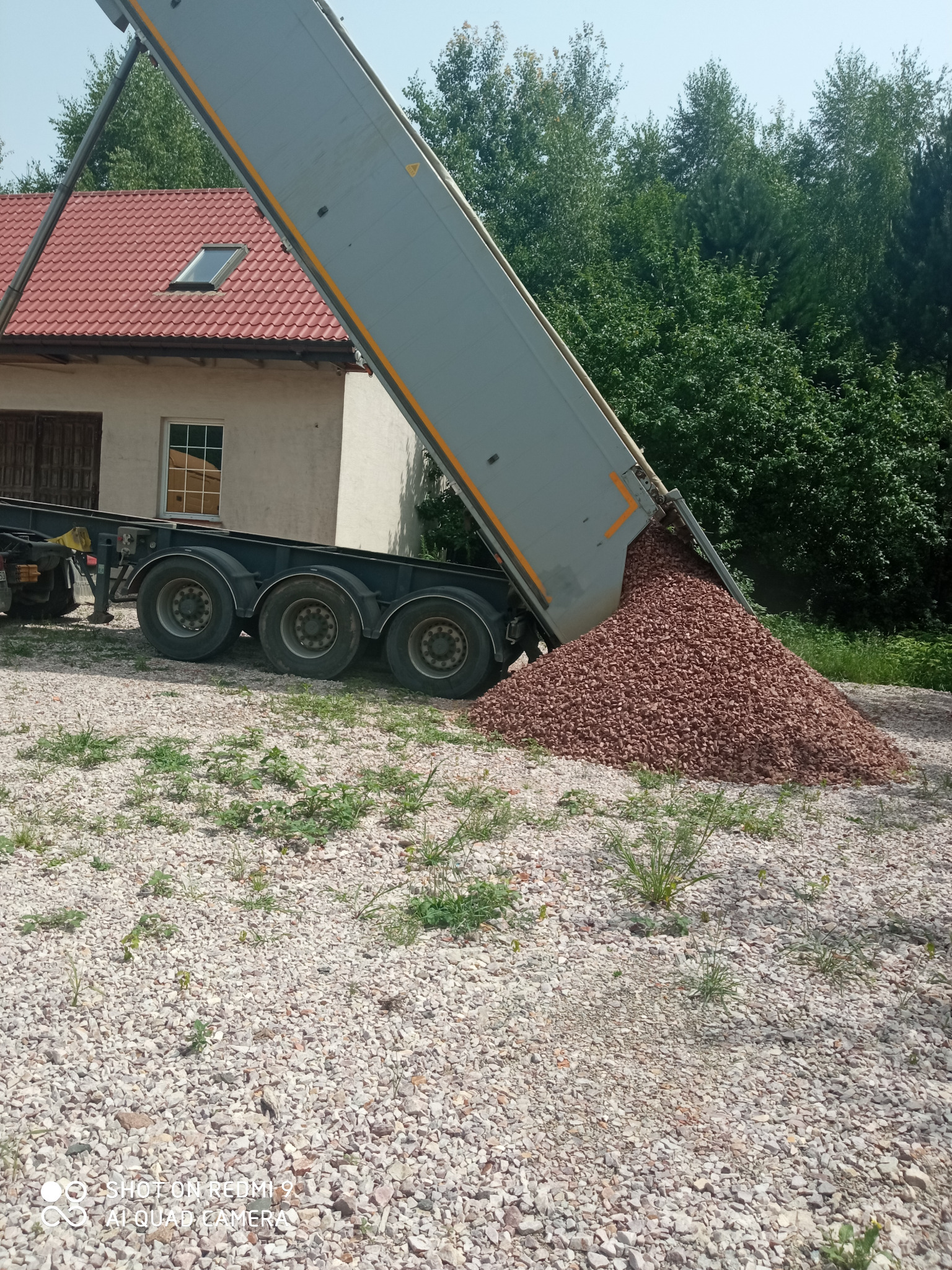 Ciężarówka z uniesioną naczepą wysypuje brązowy gruz na jasne kamienie. W tle budynek z czerwonym dachem i zieleń drzew.