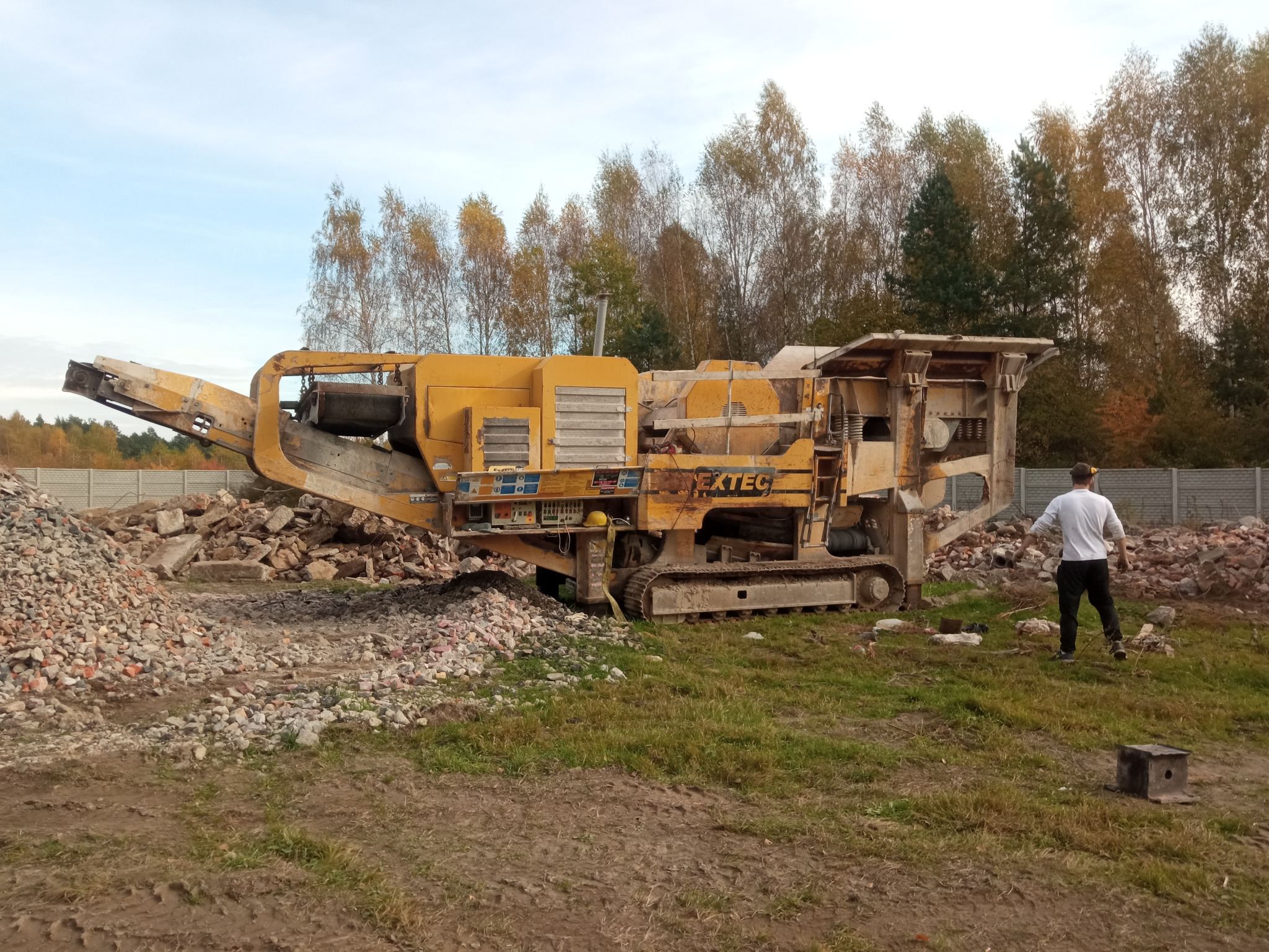 Żółty mobilny kruszarka EXTEC na gąsienicach, obok sterta gruzu. W tle drzewa i betonowe ogrodzenie. Mężczyzna w białej koszulce stoi obok maszyny.