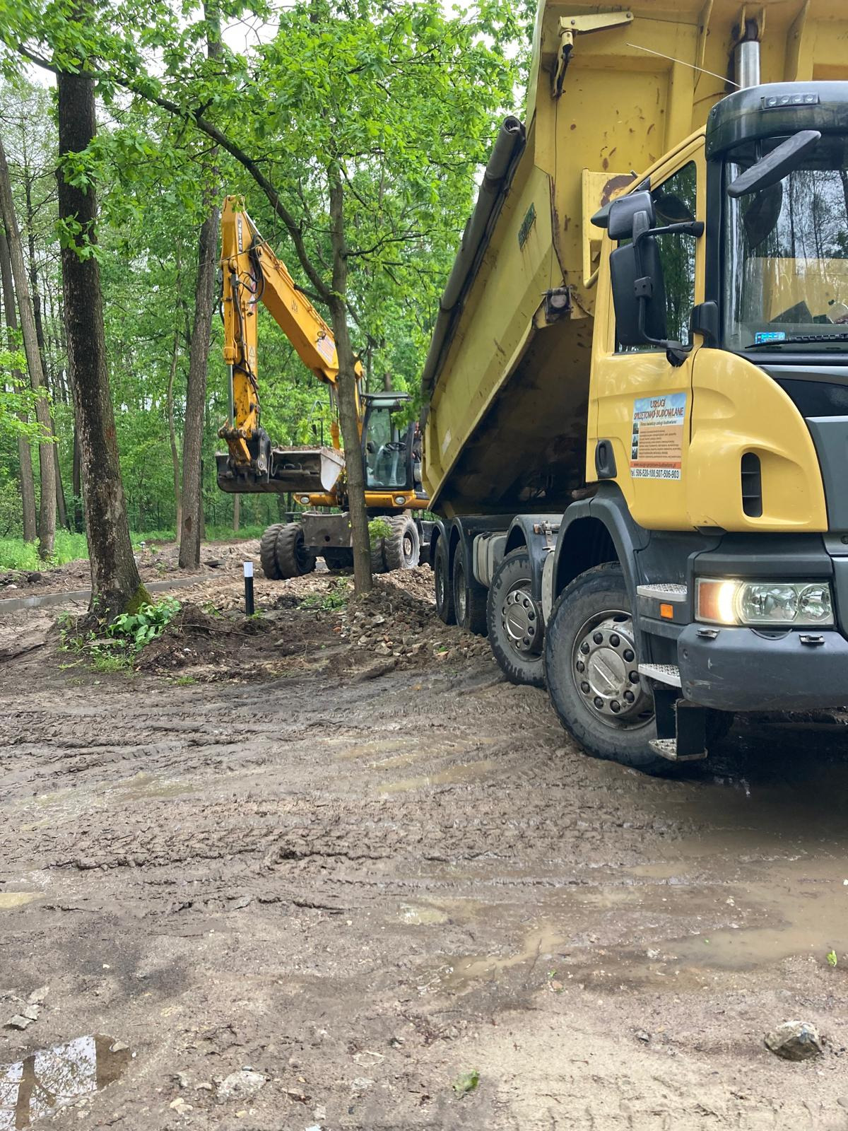 Żółta ciężarówka z podniesioną skrzynią i koparka na tle drzew, teren błotnisty. Widoczne ślady opon. Praca w toku.