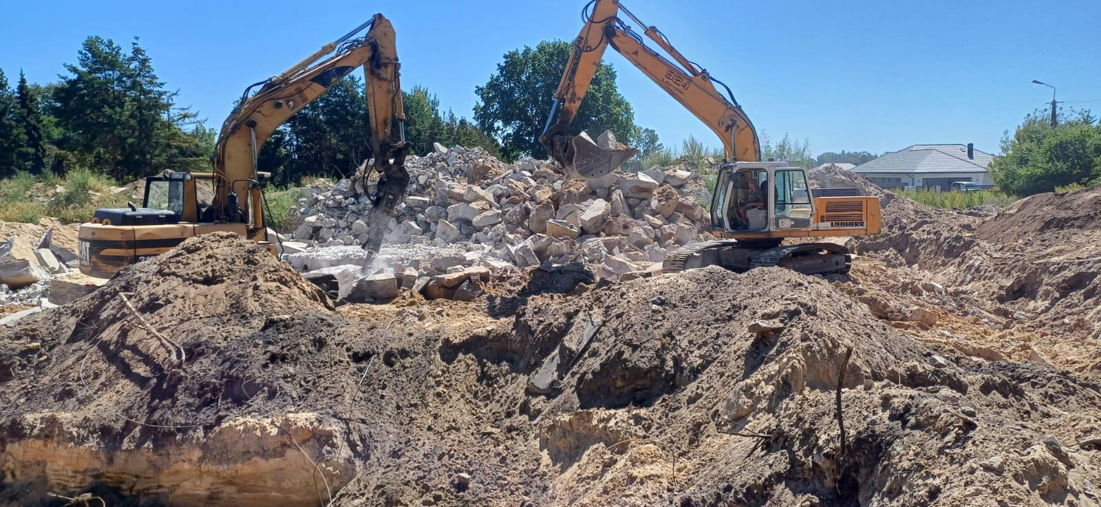Dwie żółte koparki na placu budowy, jedna kruszy gruz młotem hydraulicznym, druga nabiera gruz łyżką. W tle drzewa i dom.