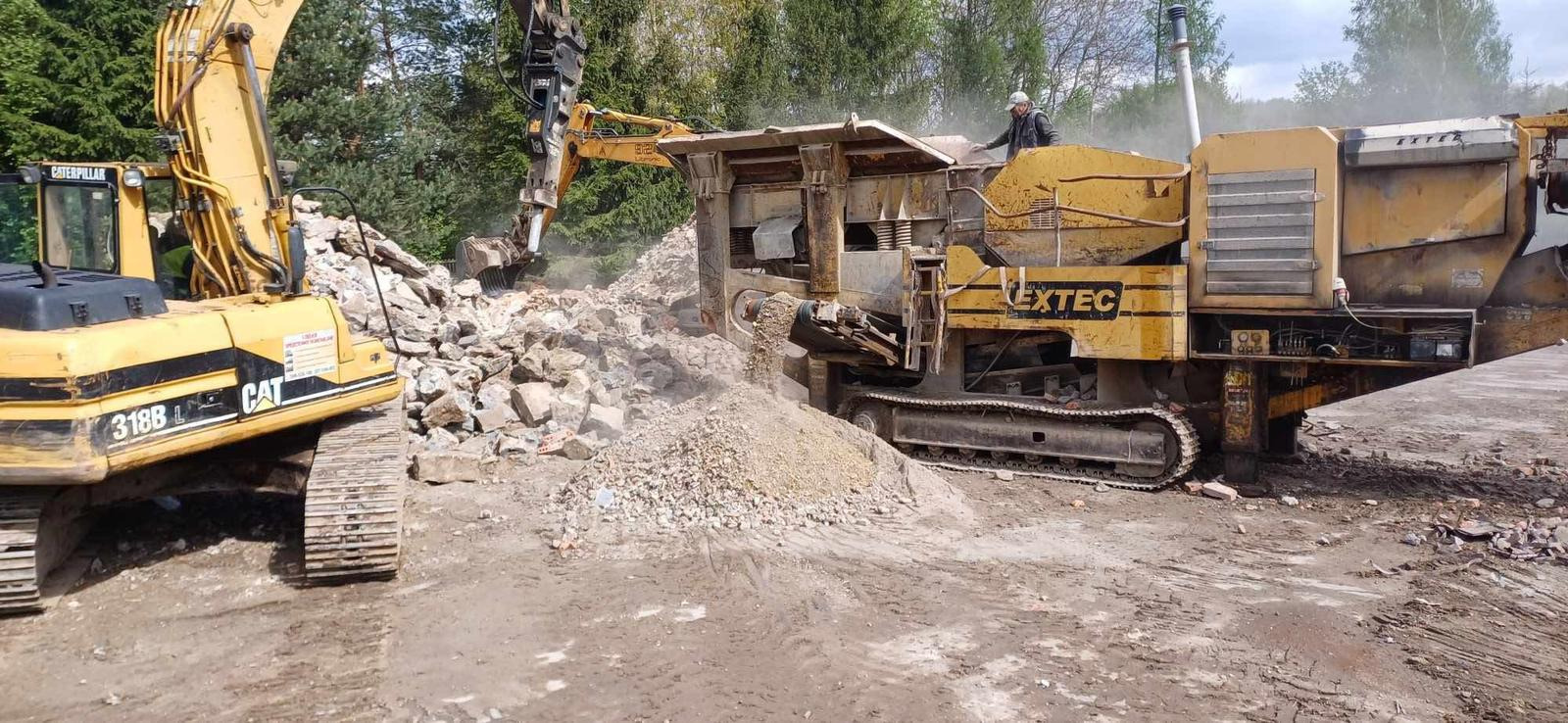Żółty sprzęt budowlany CAT 318B L i EXTEC rozdrabnia gruz na placu budowy. Operator w kabinie. Drzewa w tle.