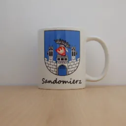 Biały kubek z herbem Sandomierza na drewnianym blacie. Herb w niebieskiej tarczy, z bramą i wieżami, zwieńczony koroną. Napis 'Sandomierz' czarną czcionką.