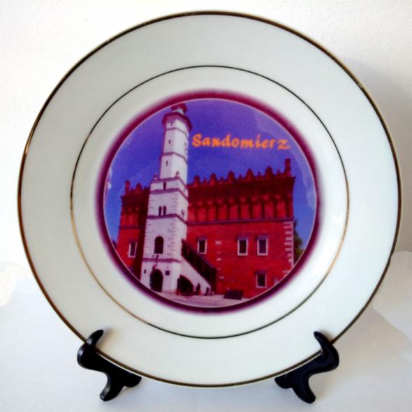 Dekoracyjny talerz z nadrukiem Ratusza w Sandomierzu, otoczony złotą obwódką, umieszczony na czarnym stojaku. Centralny kadr, jasne tło.