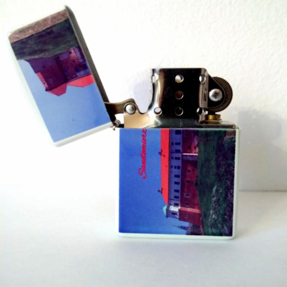 Otwarta zapalniczka z nadrukiem kolorowego zdjęcia Zamku Królewskiego w Sandomierzu, na białym tle. Widoczny mechanizm zapalniczki.