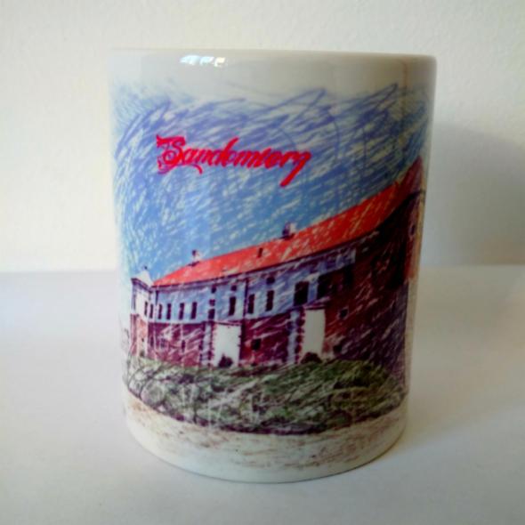 Ceramiczny kubek z ręcznie rysowanym widokiem Sandomierza, z dominującą czerwienią dachu i niebieskim niebem. Kubek stoi na białym tle.