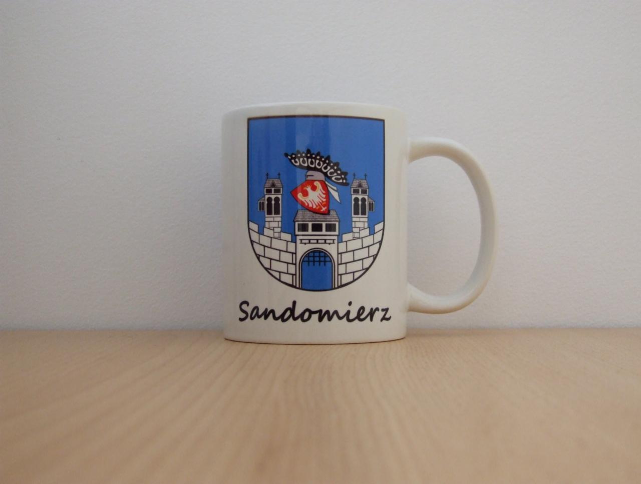 Biały kubek z herbem Sandomierza na drewnianym blacie. Herb w niebieskiej tarczy, z bramą i wieżami, zwieńczony koroną. Napis 'Sandomierz' czarną czcionką.