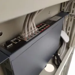 Zamontowany switch sieciowy w metalowej obudowie, widoczne kable Ethernet podłączone do portów.