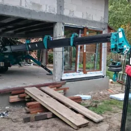 Montaż okna z użyciem dźwigu przyssawkowego, widoczni pracownicy w czerwonych rękawicach, okno z brązową ramą w surowym stanie budynku.