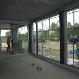 Wnętrze surowego budynku w trakcie montażu dużych okien przesuwnych z czarnymi ramami, widoczni pracownicy w kombinezonach, na zewnątrz samochody i drzewa.