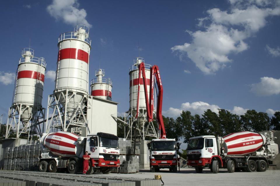 Betoniarnia z silosami i trzema betonomieszarkami z logo firmy na tle błękitnego nieba z chmurami. Na pierwszym planie ułożone płyty betonowe.