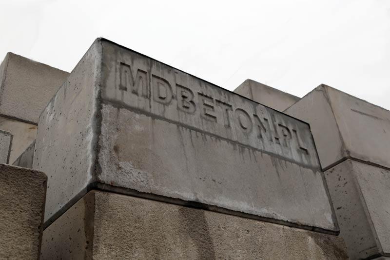Szare bloki betonowe ułożone jeden na drugim, z wyżłobionym napisem 'MDBETON.PL', widoczne z dołu pod kątem, na tle pochmurnego nieba.