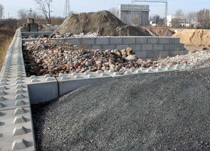 Konstrukcja oporowa z prefabrykatów betonowych, wypełniona gruzem i kamieniami, z hałdą żwiru na pierwszym planie i budynkiem w tle.