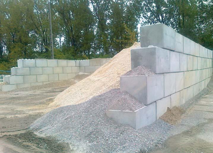 Ściana oporowa z betonu modułowego, z widocznymi blokami w kształcie klocków Lego, usypana ziemią i żwirem, na tle zieleni. Elementy prefabrykowane.