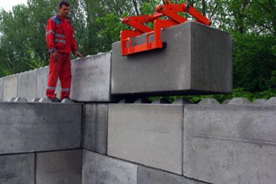 Montaż prefabrykowanych bloków betonowych za pomocą dźwigu; pracownik w czerwonym kombinezonie nadzoruje proces budowy ściany z szarych elementów.