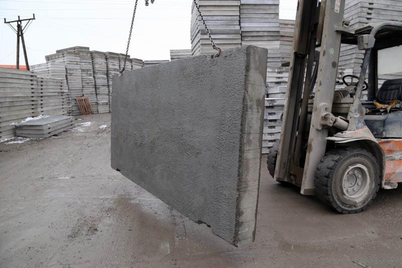 Płyta betonowa o szorstkiej fakturze zawieszona na łańcuchach, w tle stosy płyt i wózek widłowy.