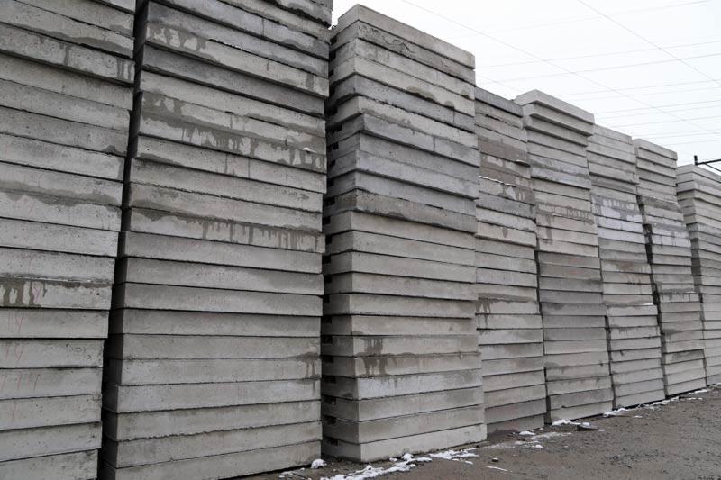 Stosy szarych płyt betonowych na placu składowym, widoczne ślady po wodzie, niebo w tle. Płyty ułożone równo, tworząc geometryczny wzór.