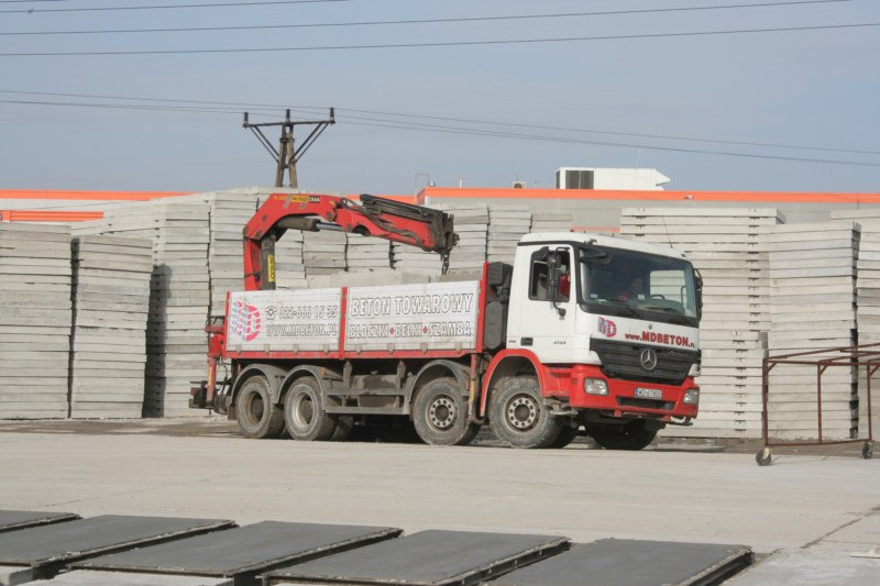 Ciężarówka z dźwigiem na placu, obok stosy szarych płyt betonowych. Na boku ciężarówki reklama betonu towarowego.