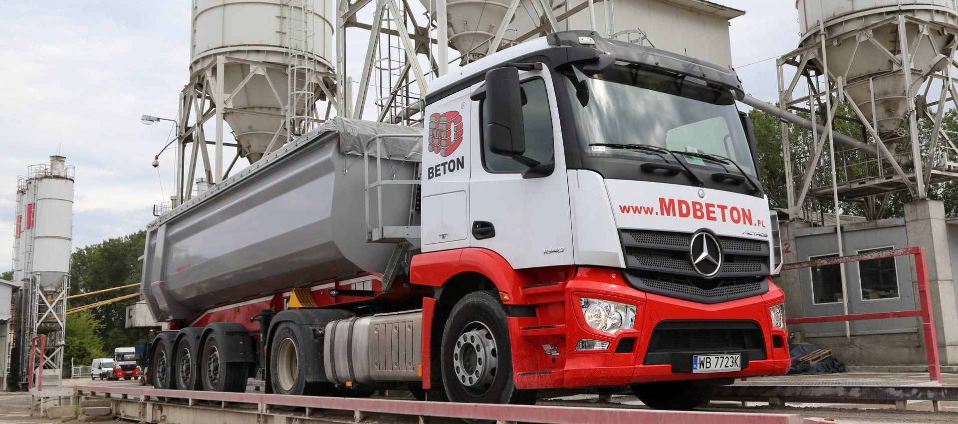 Ciężarówka z naczepą do transportu betonu, oznaczona logo i adresem strony internetowej, stoi na wadze na tle silosów betoniarni.