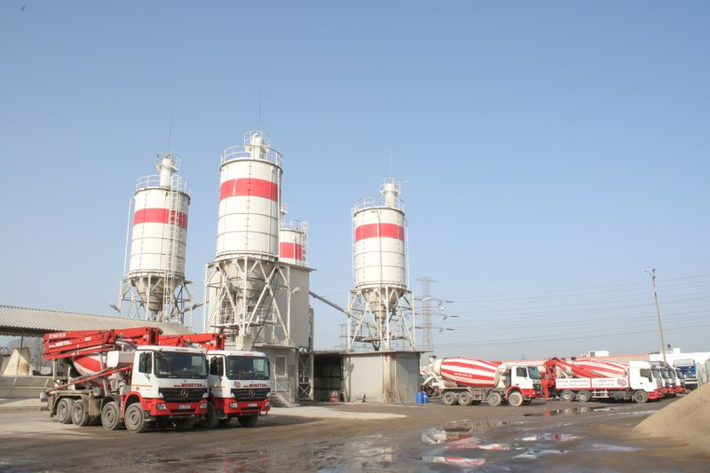 Widok na teren zakładu z silosami i betonomieszarkami w barwach biało-czerwonych, z widocznym logo 'Mdbeton' na kabinach pojazdów.