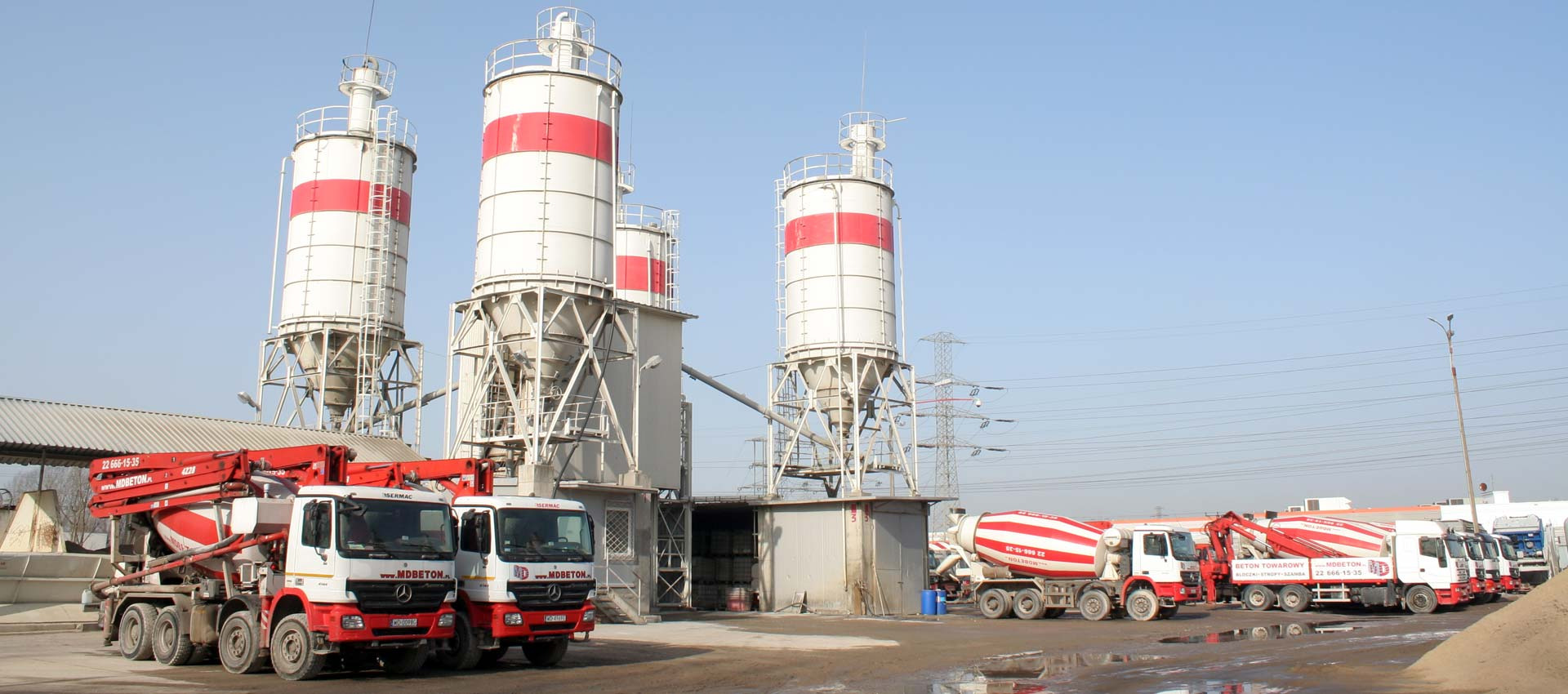 Widok na betoniarnię z wysokimi silosami w biało-czerwone pasy i zaparkowanymi betonomieszarkami z logo firmy na tle błękitnego nieba.