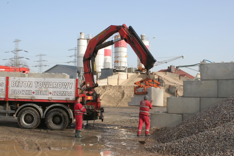 Ciężarówka z napisem 'Beton Towarowy' i dźwigiem przenosi betonowy prefabrykat na placu budowy; w tle silosy betoniarni i hałda piasku.