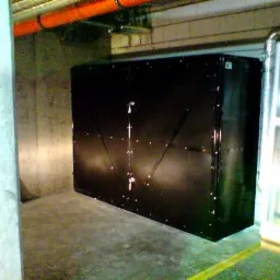 Czarna, metalowa szafa z widocznymi nitami i wzmocnieniami w garażu podziemnym. Pomarańczowe rury pod sufitem, surowy beton na ścianach.