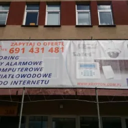 Baner firmy Krypton Telesystemy na budynku, oferującej monitoring, systemy alarmowe, sieci komputerowe, światłowodowe i dostęp do internetu. Widoczne logo BCS Satel i Facebook.