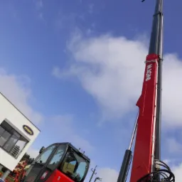 Czerwona ładowarka teleskopowa MAGNI z wysuniętym ramieniem na tle błękitnego nieba. Widoczna kabina operatora i logo firmy RIVAL na budynku w tle.