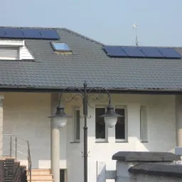 Dom z szarym dachem i panelami słonecznymi. Widoczny ozdobny słup oświetleniowy przed budynkiem i okno dachowe. Jasna elewacja i ciemna dachówka.