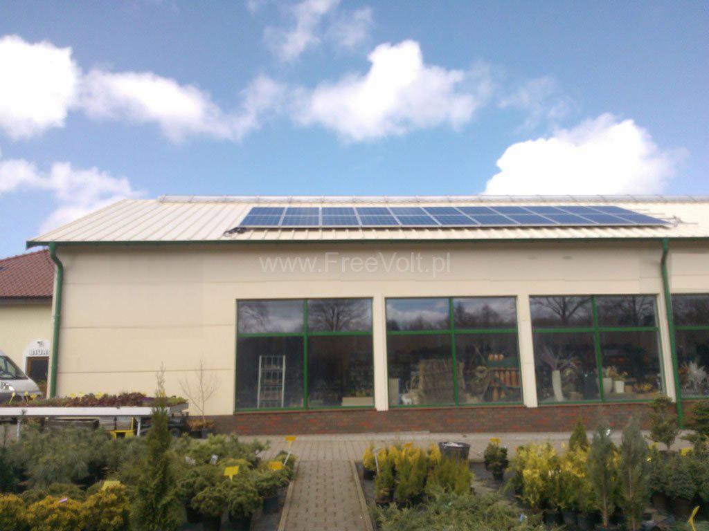 Biały budynek z panelami słonecznymi na dachu, widoczne duże okna i rośliny przed budynkiem. Na budynku widoczny adres strony internetowej.