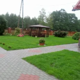 Zadbany trawnik z drewnianą altaną i brukowanym chodnikiem, widok z boku budynku z czerwonej cegły.