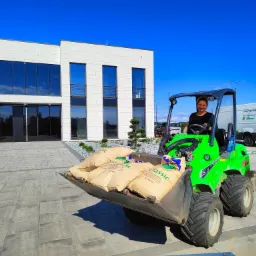 Kobieta obsługuje zieloną miniładowarkę Avant 630 z workami nasion trawy Classic na tle nowoczesnego budynku biurowego i zaparkowanego busa z logo firmy ogrodniczej.