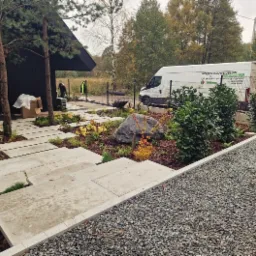 Nowoczesny ogród z betonowymi płytami chodnikowymi i ozdobnym kamieniem, świeżo nasadzone rośliny na korze, w tle czarny dom i bus firmy ogrodniczej.