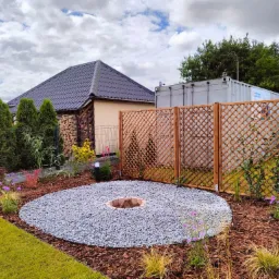 Ogród z okrągłym paleniskiem wyłożonym szarym żwirem, otoczonym korą i niską roślinnością, z drewnianą kratownicą w tle.