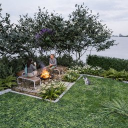 Ogrody Ślusarczyk - Ogród z paleniskiem otoczonym kamienną nawierzchnią, ławkami i roślinnością. W tle widoczne drzewa, krzewy, kwiaty, trawnik i robot koszący. Siedzące osoby przy ogniu.