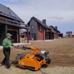 Ogrody Ślusarczyk - Mężczyzna obsługuje pomarańczową maszynę do przygotowania gruntu pod trawnik na tle nowoczesnych domów z cegły i wyrównanej ziemi.