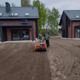 Ogrody Ślusarczyk - Przygotowanie terenu pod trawnik: glebogryzarka spalinowa w akcji na działce z dwoma nowoczesnymi domami z cegły. Widoczne ślady opon i pracownicy w trakcie pracy.