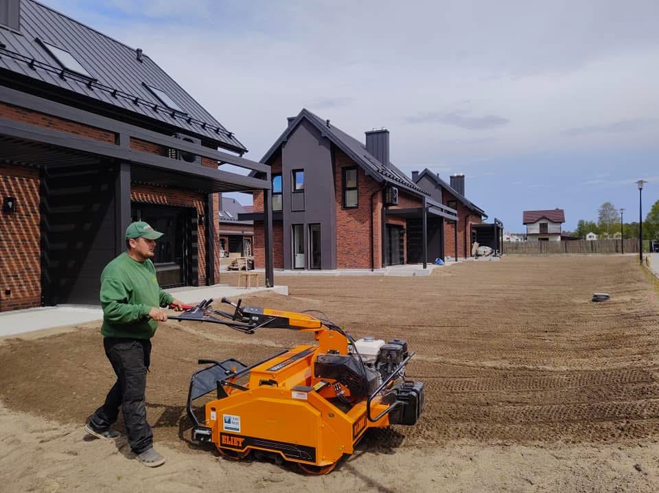 Mężczyzna obsługuje pomarańczową maszynę do przygotowania gruntu pod trawnik na tle nowoczesnych domów z cegły i wyrównanej ziemi.
