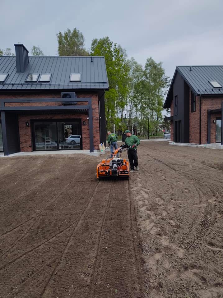 Przygotowanie terenu pod trawnik: glebogryzarka spalinowa w akcji na działce z dwoma nowoczesnymi domami z cegły. Widoczne ślady opon i pracownicy w trakcie pracy.