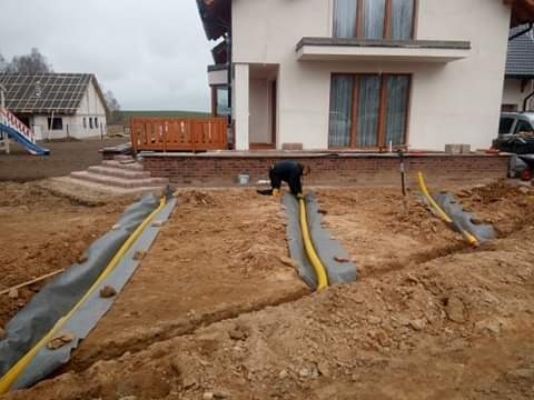 Instalacja drenażu: żółte rury w wykopach wyłożonych geowłókniną, pracownik w trakcie montażu przy fundamencie domu, widoczne schody i taras.